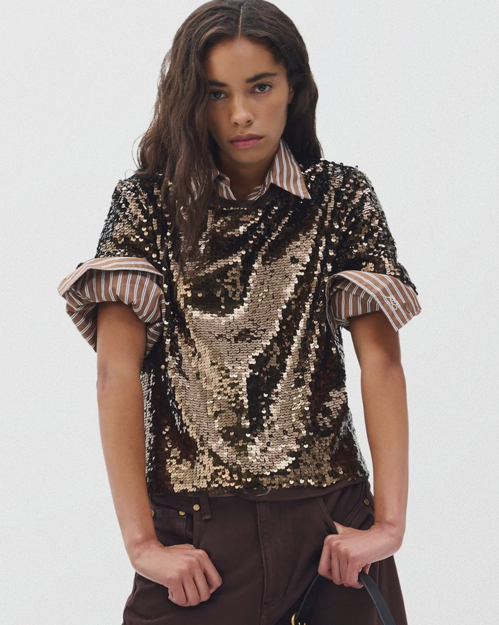 A woman with wavy brown hair wears the Rag & Bone lucy sequin tee—a relaxed fit black and gold sequin top—layered over a brown and white striped shirt with rolled sleeves, paired with brown pants against a plain light background.