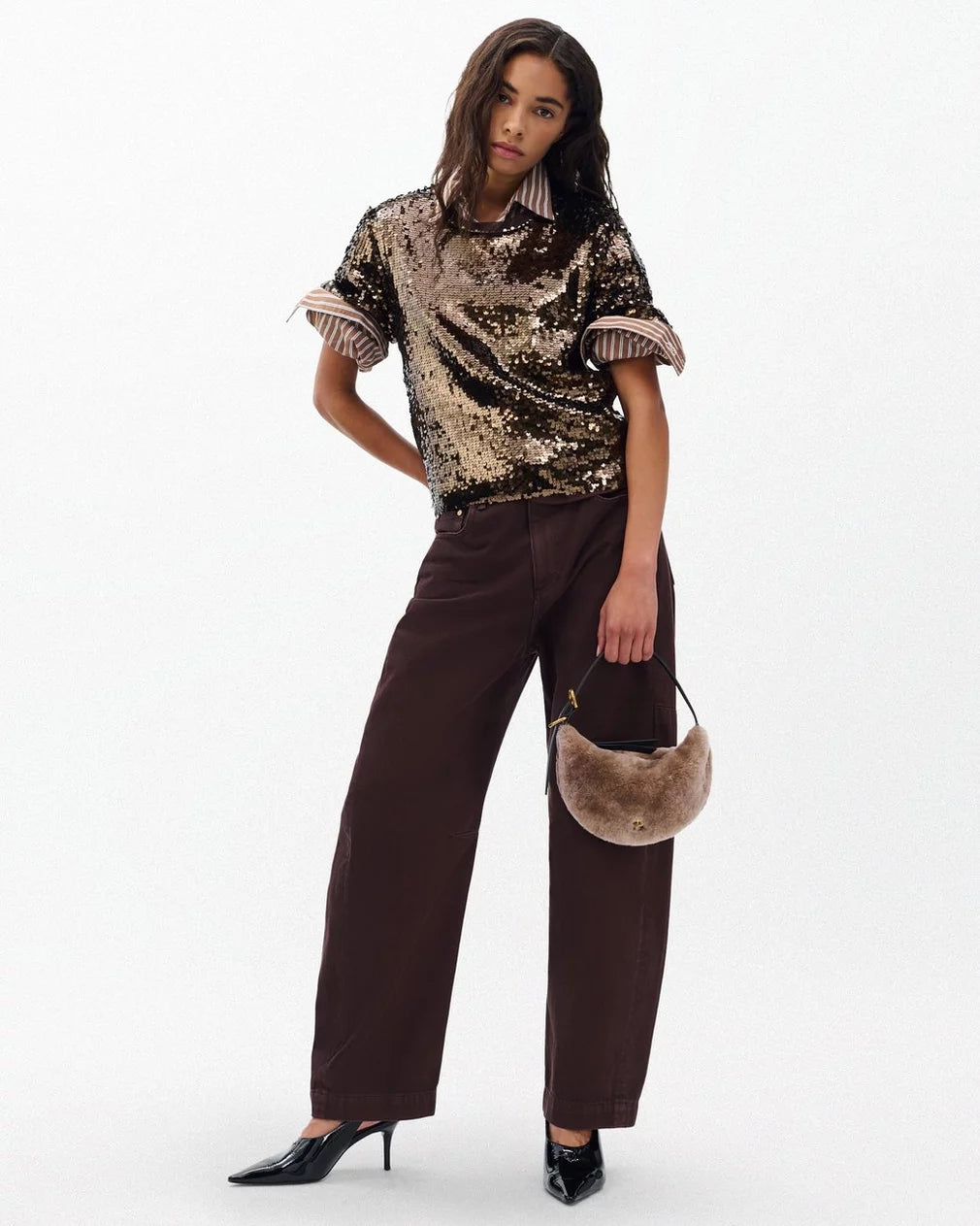 A woman stands against a plain background wearing the Rag & Bone lucy sequin tee, wide-leg brown pants, black heeled shoes, and holding a small round beige fur handbag—an ideal look for party season.