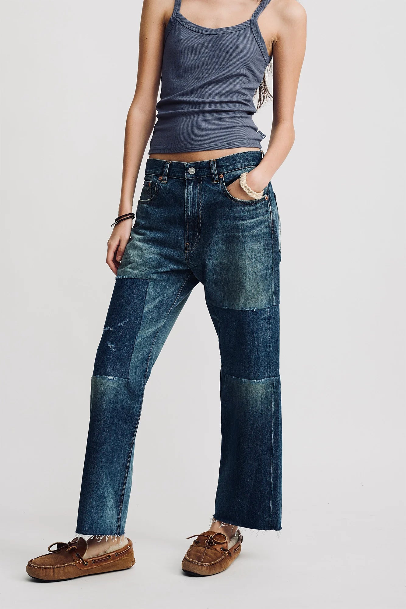 A person stands against a plain light background, hands in pockets, wearing a fitted gray tank top, Denimist Lucy Boyfriend Jeans with patchwork and frayed hems, and brown moccasin-style shoes.