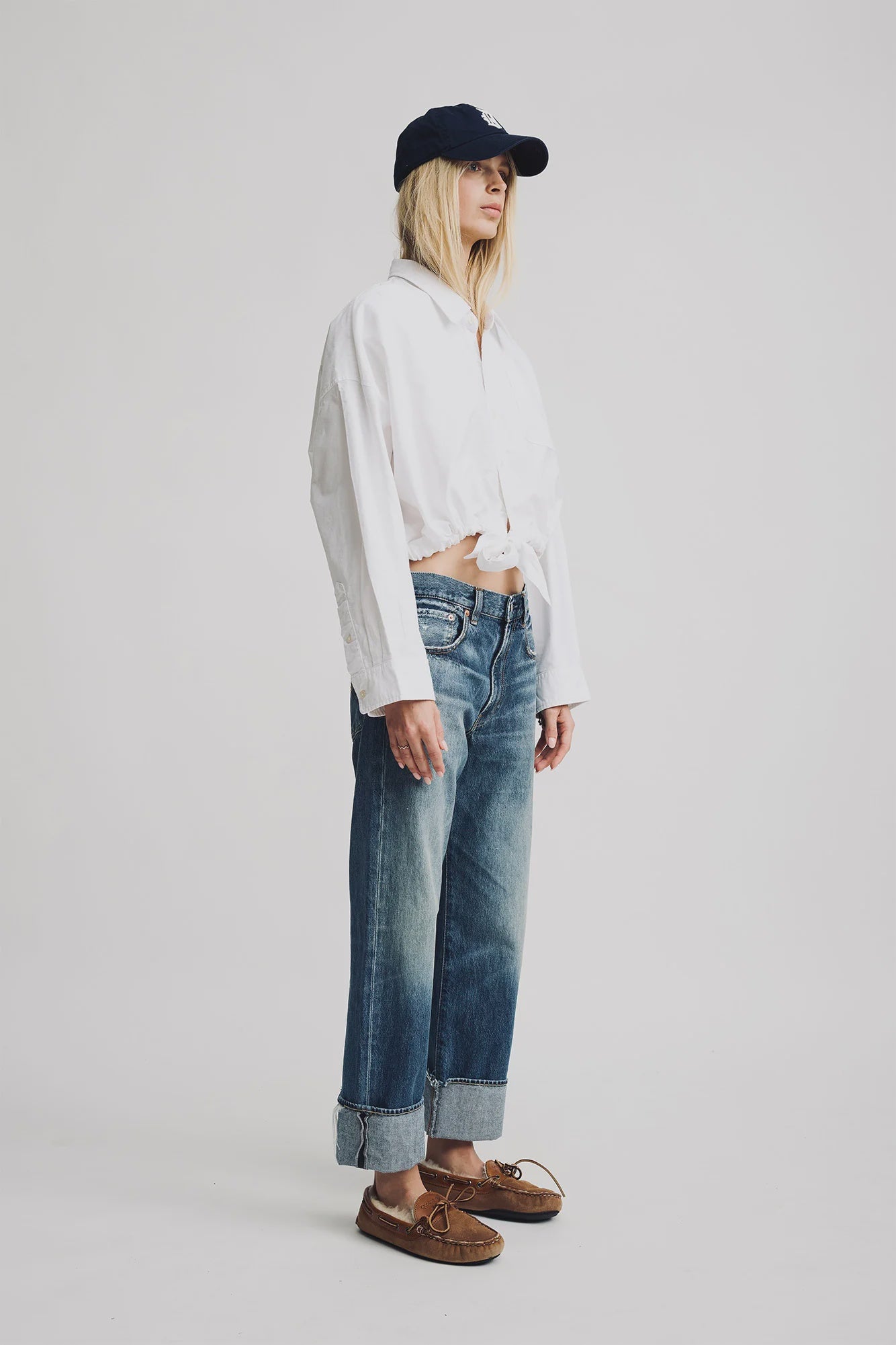 A person stands against a plain background wearing a navy baseball cap, a loose white cropped long-sleeve shirt, Denimist Lucy Boyfriend Jeans in 100% cotton with cuffs, and brown boat shoes showing part of the midriff.