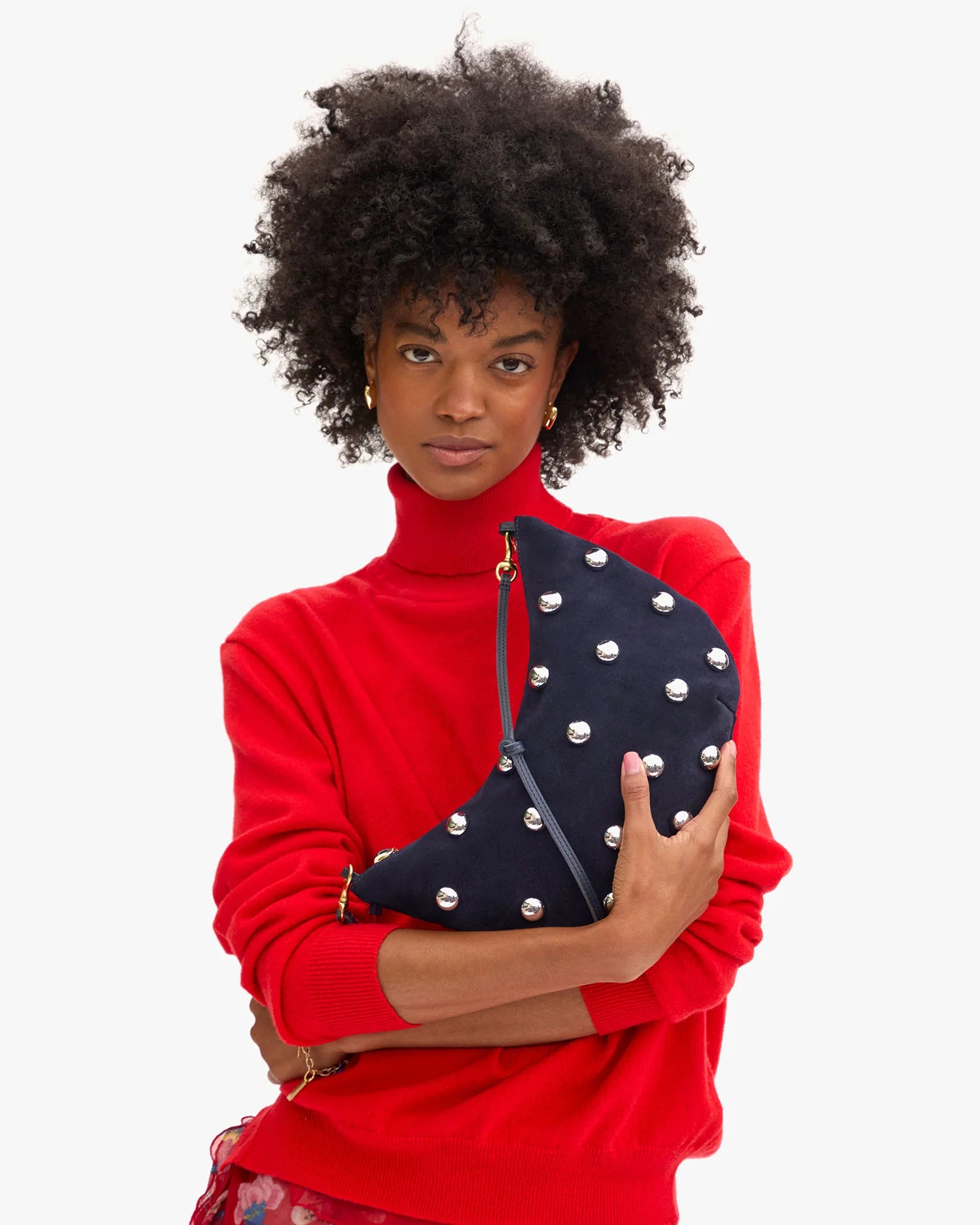 A woman with curly hair in a bright red turtleneck holds the Clare Vivier F25- Lune, a black crescent-shaped handbag with silver studs and a knotted strap, standing against a plain white background.