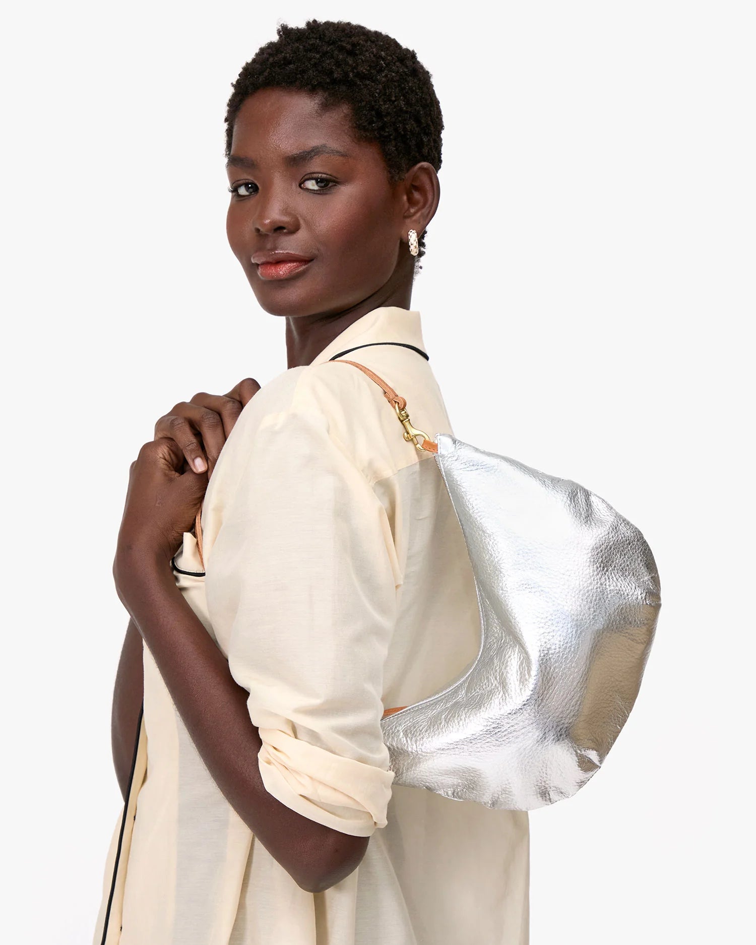 A woman in a light-colored shirt stands with her back slightly turned, looking at the camera and holding the Clare Vivier Lune, a metallic silver chrome-tanned leather shoulder bag with a brown strap.