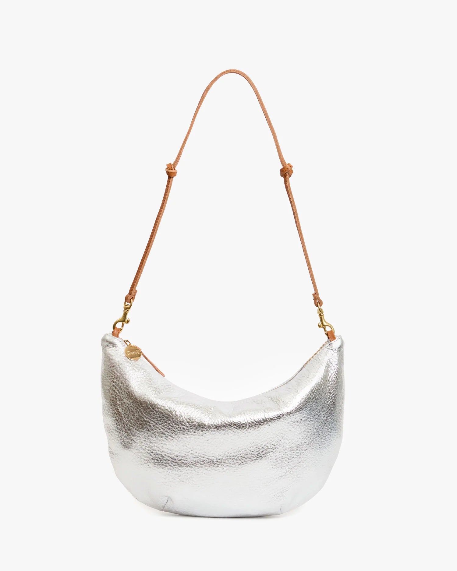 The Clare Vivier Lune is a crescent-shaped metallic silver handbag featuring a textured surface and a brown chrome-tanned leather shoulder strap, displayed against a plain white background.