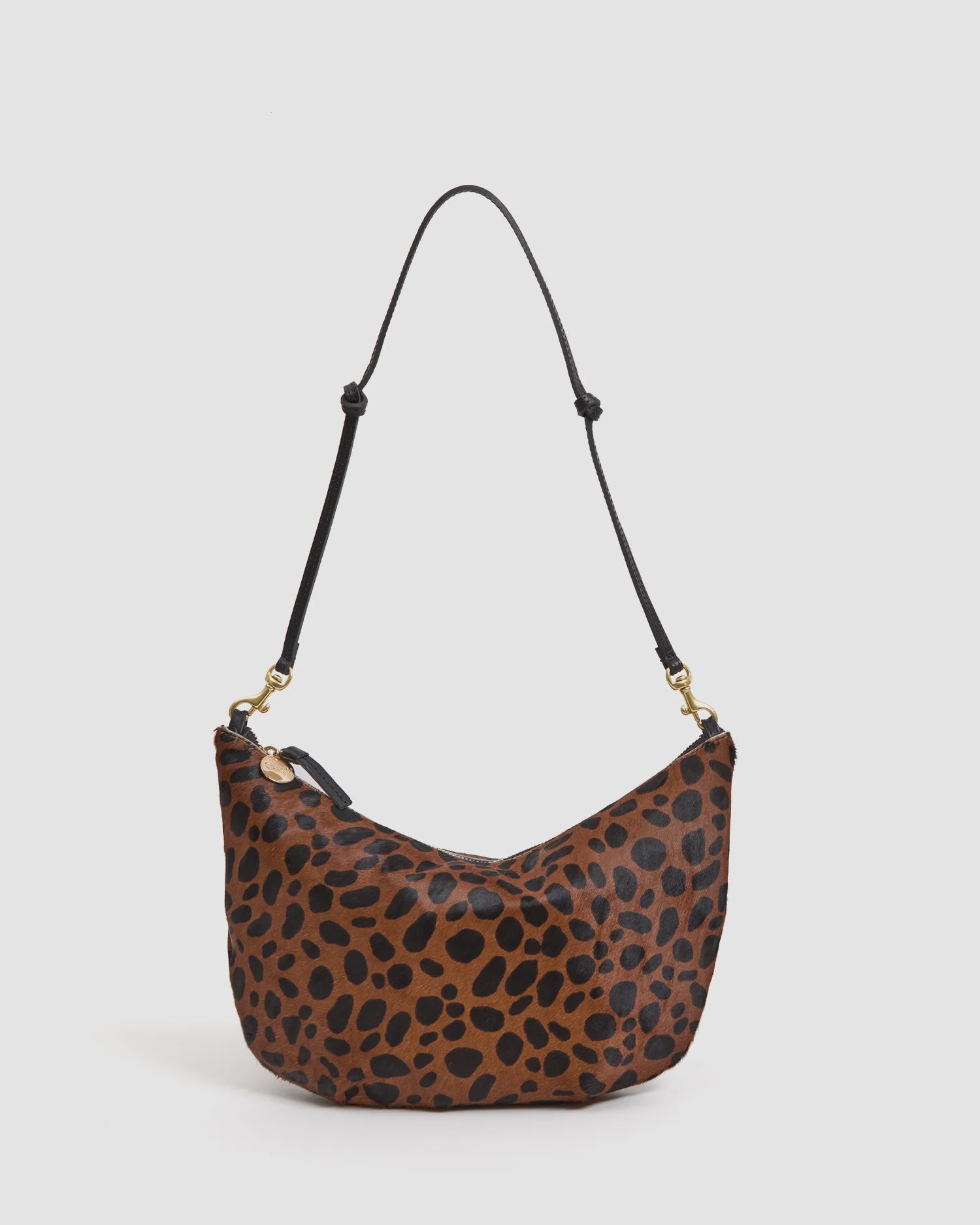 The F25- Lune by Clare Vivier is a curved brown shoulder bag in black leopard-print hair-on leather, featuring a zipper closure and an adjustable black strap with gold hardware.