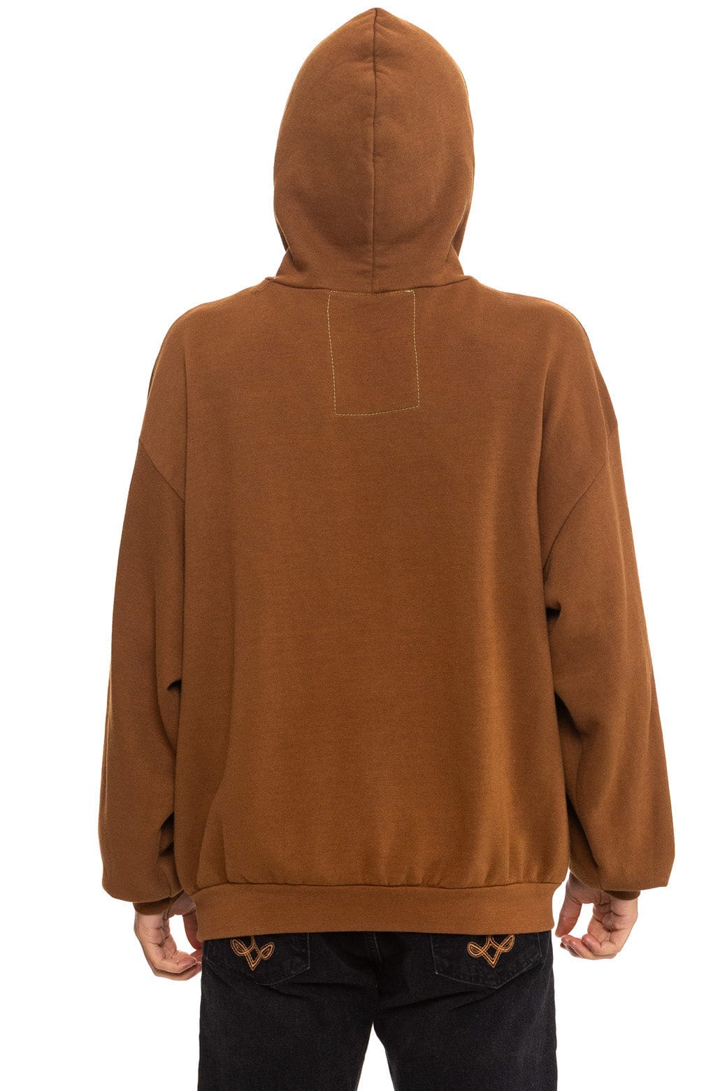 Someone wearing the Aviator Nation "locals only - pullover hoodie relaxed" in brown with the hood up, seen from behind. The unisex, loose-fit hoodie is paired with black pants featuring small embroidered designs on the back pockets.