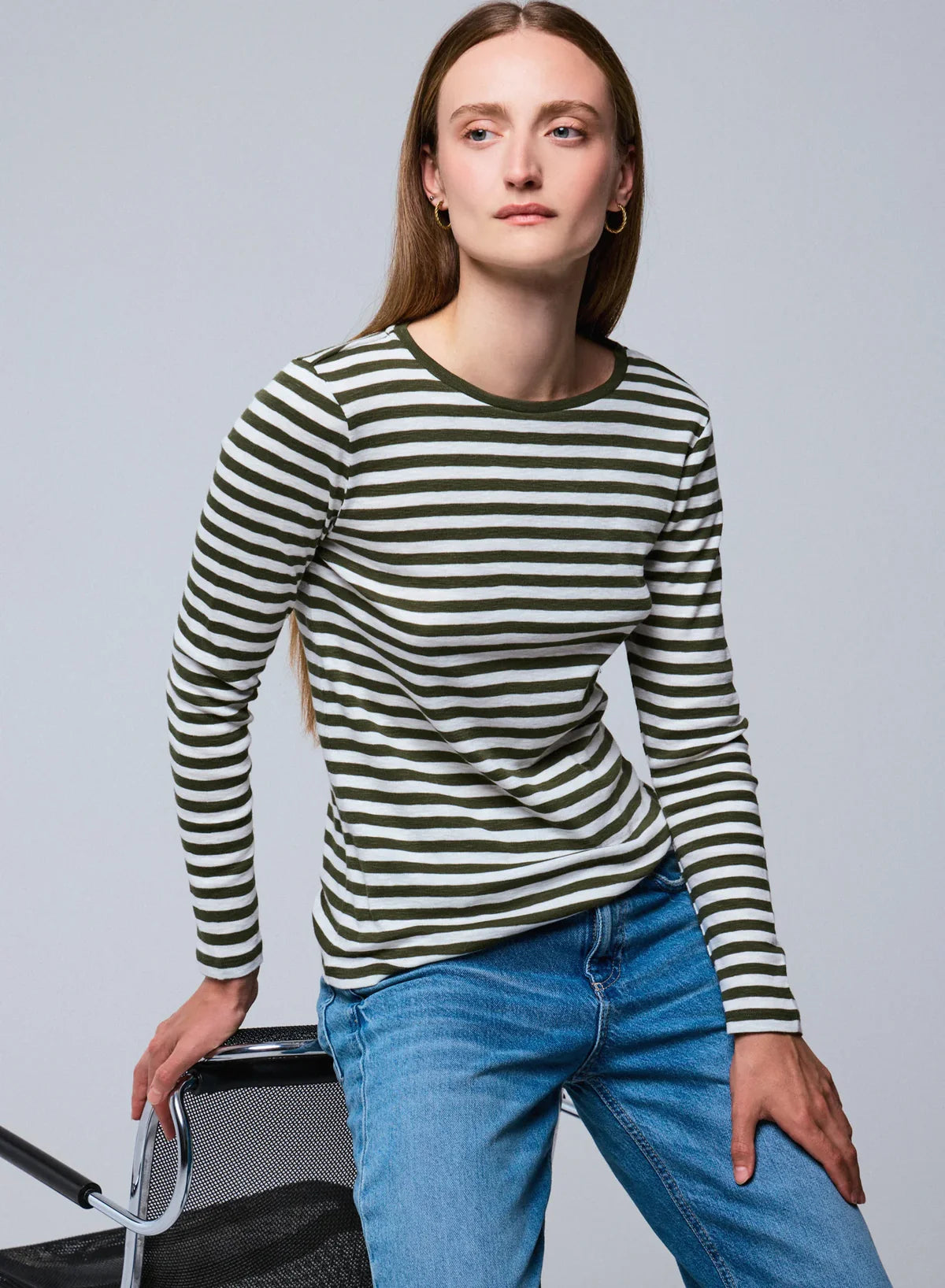 A woman with straight brown hair wears the Majestic Filatures Organic Cotton Stripe L/S Crewneck in green and white stripes with blue jeans, seated on a black chair against a light grey background.