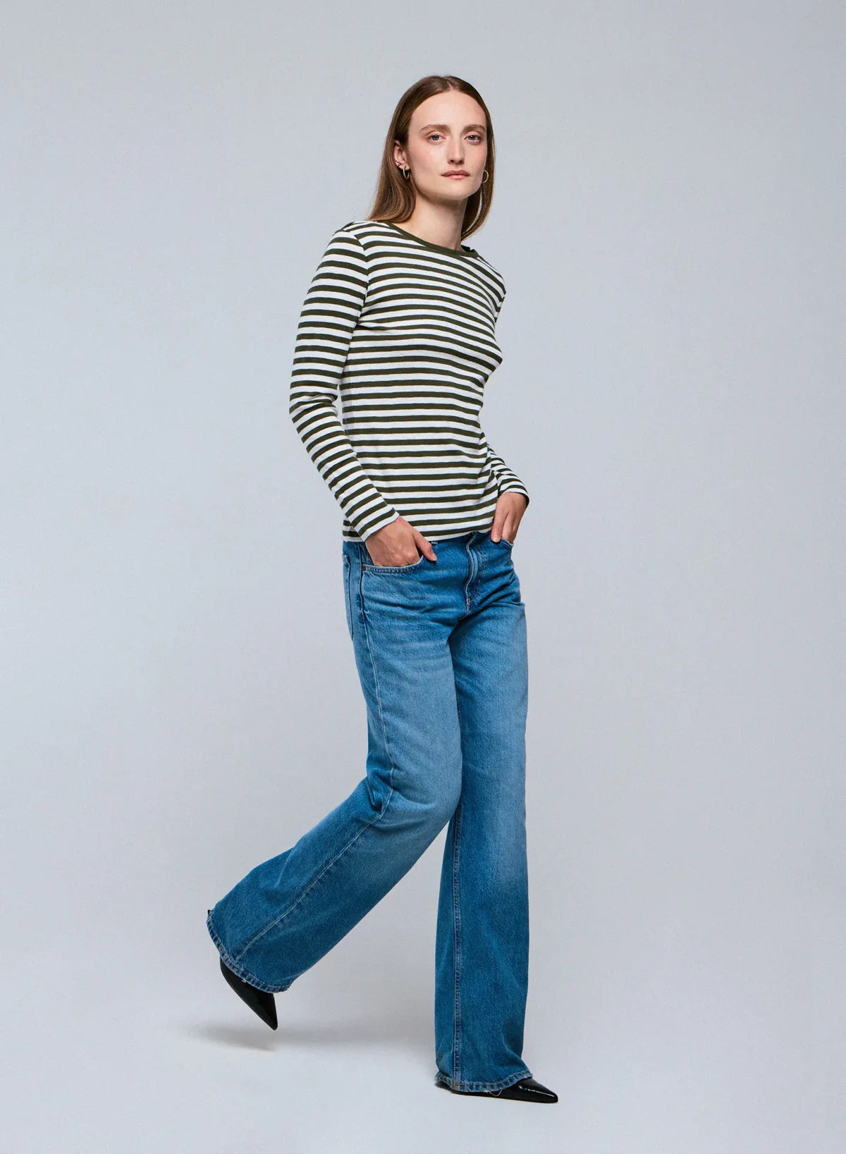 A woman stands with her hands in her jeans pockets, wearing the Majestic Filatures Organic Cotton Stripe L/S Crewneck and wide-leg blue jeans against a plain light gray background.