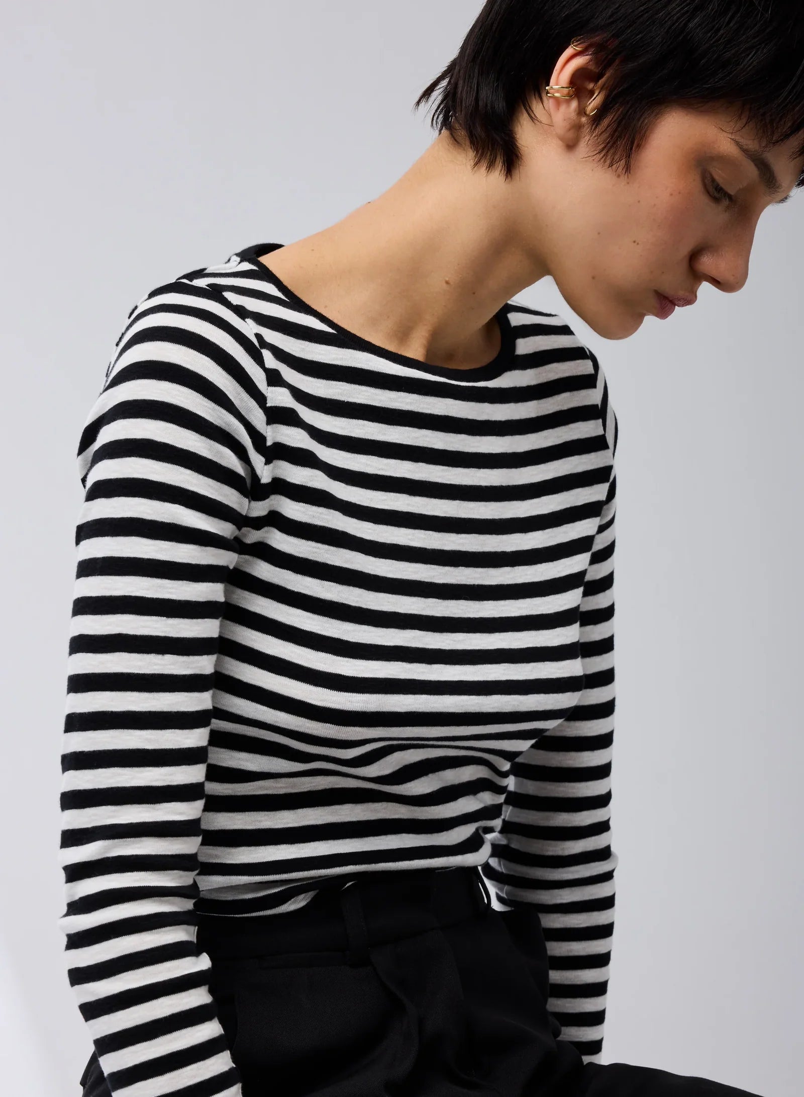 A person with short dark hair is seated against a plain light background, wearing the Majestic Filatures Organic Cotton Stripe L/S Crewneck and black pants.