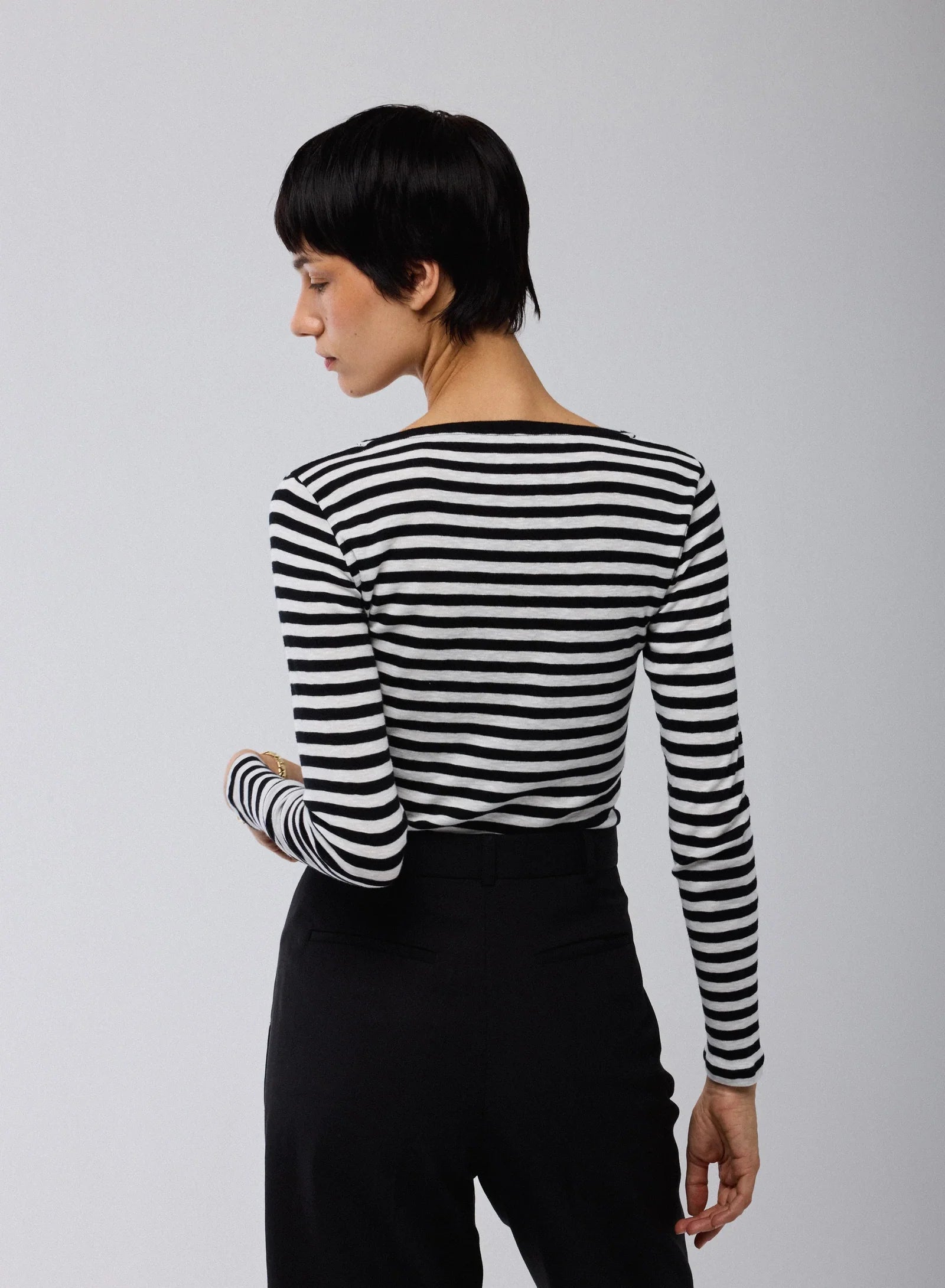A person with short dark hair faces away, wearing Majestic Filatures’ Organic Cotton Stripe L/S Crewneck in black and white stripes with black pants, set against a plain light gray background.