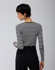 A person with short dark hair faces away, wearing Majestic Filatures’ Organic Cotton Stripe L/S Crewneck in black and white stripes with black pants, set against a plain light gray background.