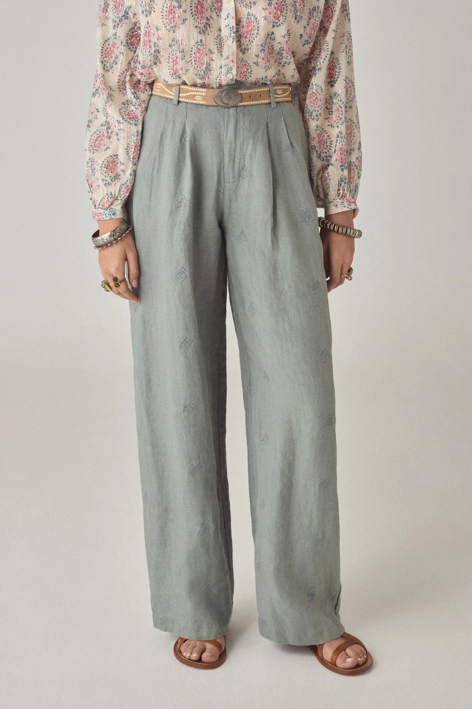 A person wearing Maison Hotel's Otto Long Pant, a patterned long-sleeve blouse, a woven belt, brown sandals, and layered jewelry stands against a plain background.