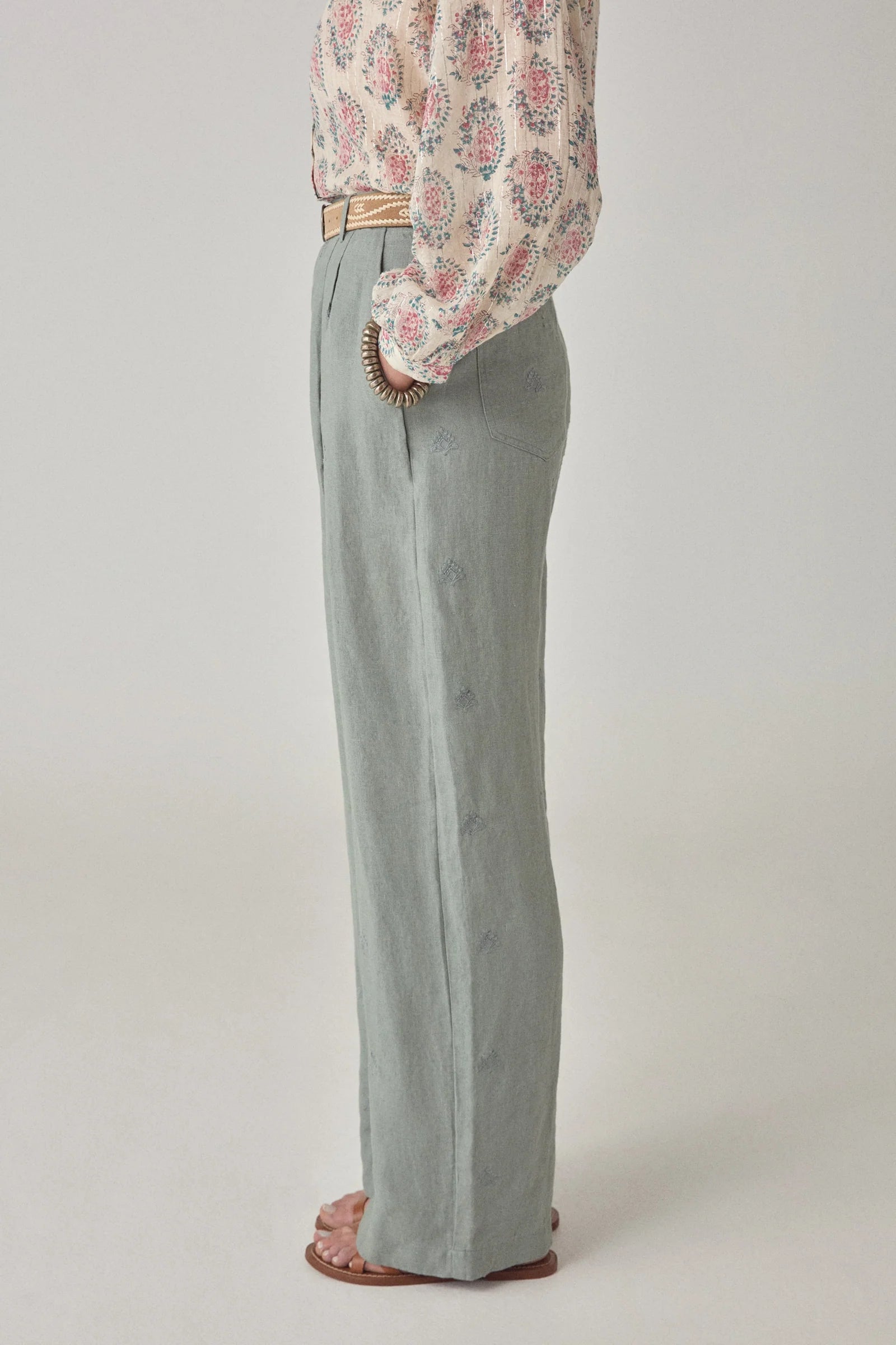A person in profile wears Maison Hotel’s Otto Long Pant in pale dusty blue, paired with a floral blouse, beige belt, and brown sandals against a plain light background.