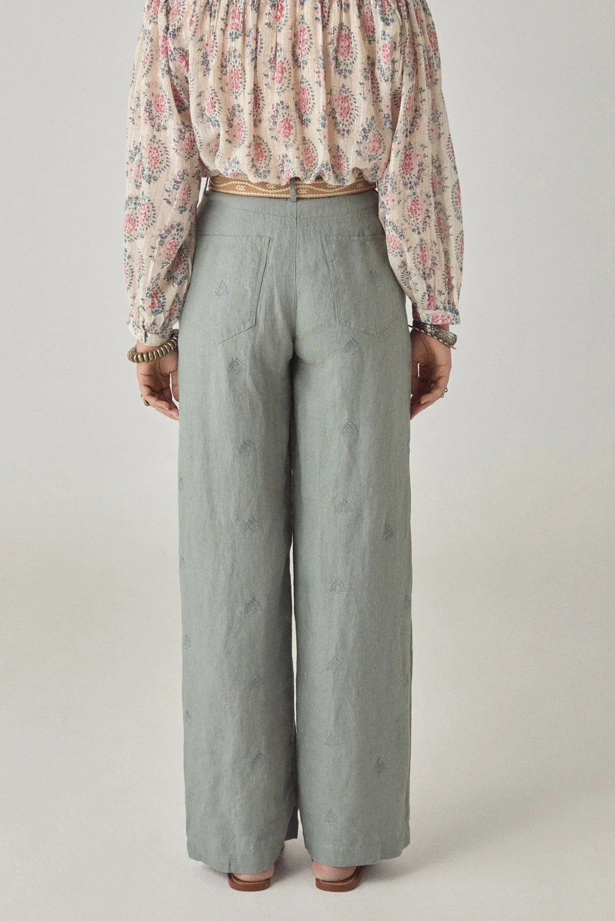 A person stands facing away in Maison Hotel's Otto Long Pant, wide-leg and pale dusty blue, paired with a tucked floral blouse, brown sandals, and bracelets. The plain background keeps the look minimal and stylish.