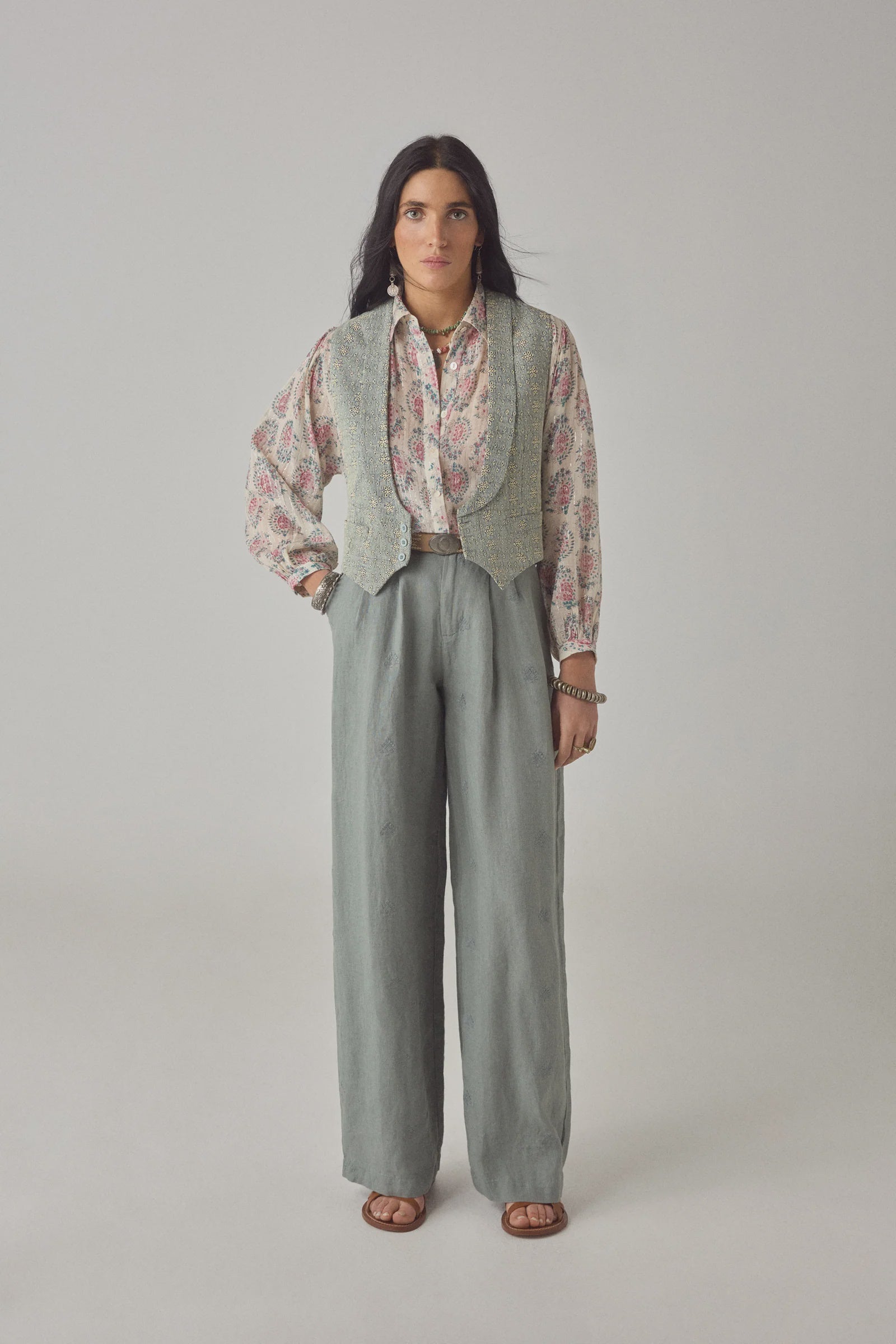 A person stands facing forward against a plain light background, wearing the Otto Long Pant by Maison Hotel, styled with a floral button-up shirt, gray textured vest, brown sandals, and jewelry.