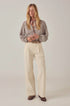 A woman stands against a plain background, wearing Maison Hotel's Theodore Pant in cream embroidered cotton twill with an eyelet pattern, a checked long-sleeve blouse, brown shoes, and a patterned neck scarf. Her hands are clasped and her blond hair is loose.