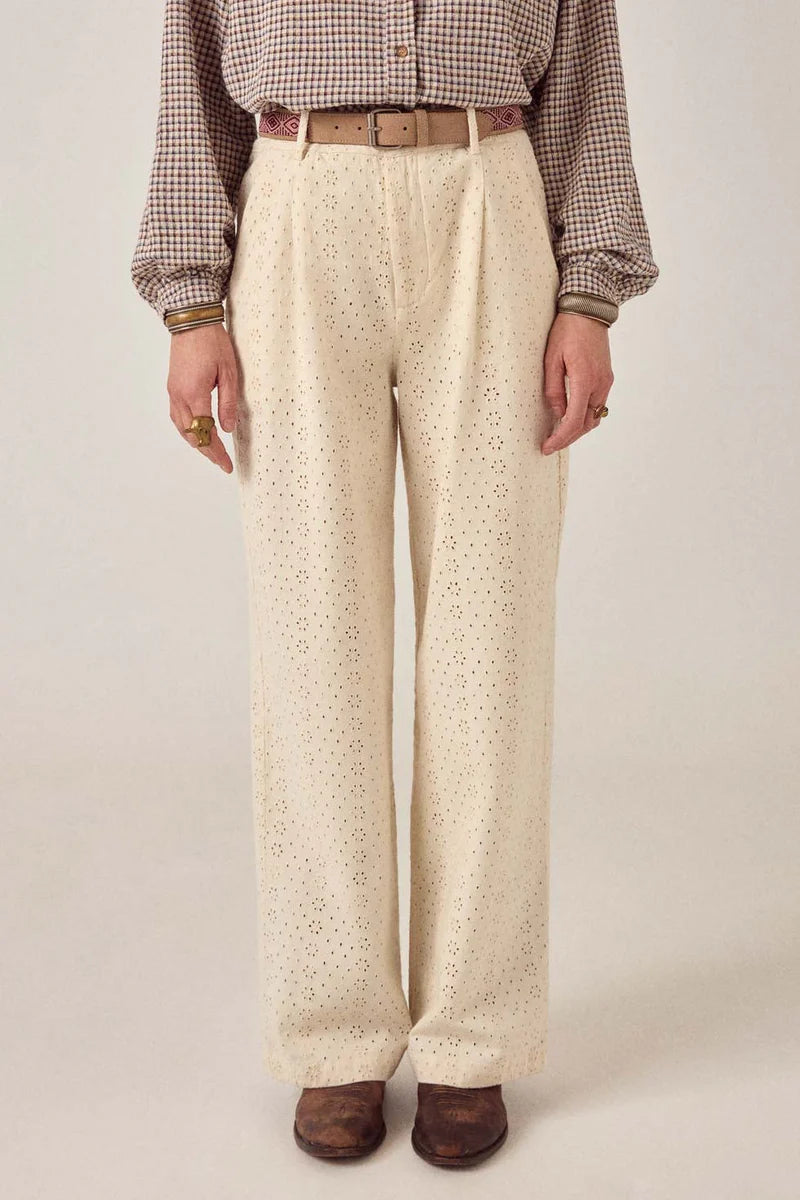 A person wears Maison Hotel's Theodore Pant—high-waisted, wide-leg, cream eyelet-embroidered cotton twill—styled with a tucked-in plaid shirt, beige belt, and brown shoes. Only the body from the shoulders down is visible against a neutral background.