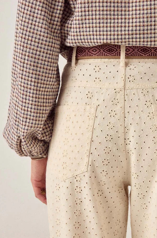 Seen from the waist down and behind, a person wears Maison Hotel's Theodore Pant—cream high-waisted wide-leg trousers in embroidered cotton twill with a floral eyelet pattern—paired with a checked shirt and burgundy patterned belt.