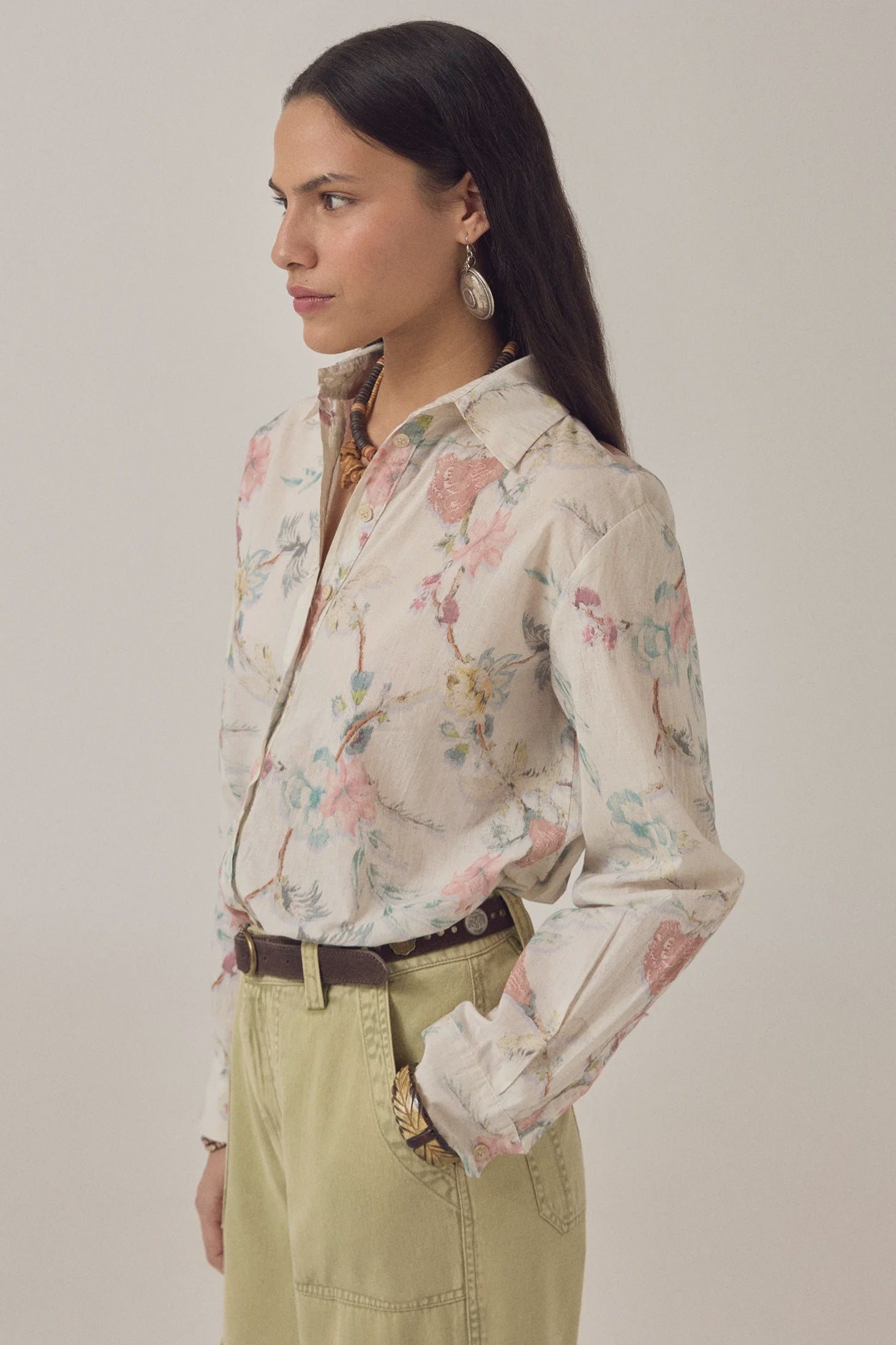 A woman with long dark hair wears the Narciso Shirt by Maison Hotel, an oversized cotton floral print top tucked into high-waisted khaki pants with a brown belt, earrings, and a patterned scarf. She stands in profile against a plain background.