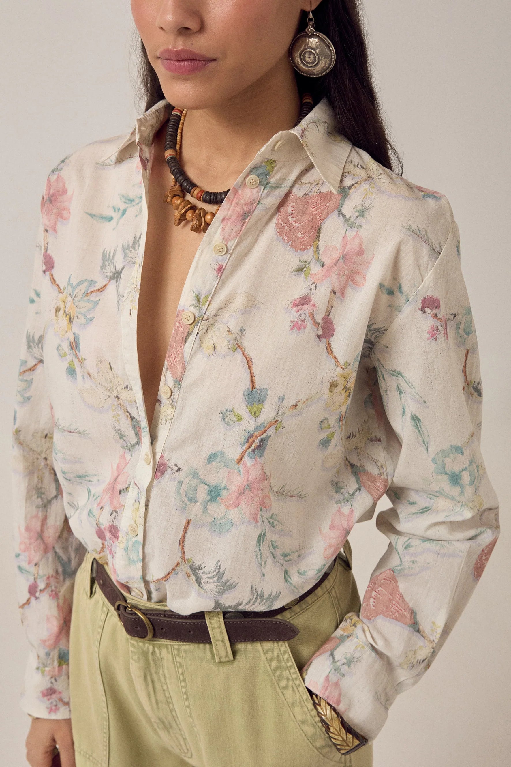A woman wears the Narciso Shirt by Maison Hotel, a floral print top tucked into high-waisted beige pants with a dark belt. She accessorizes with a necklace and large round earring; only her head and upper body are visible.