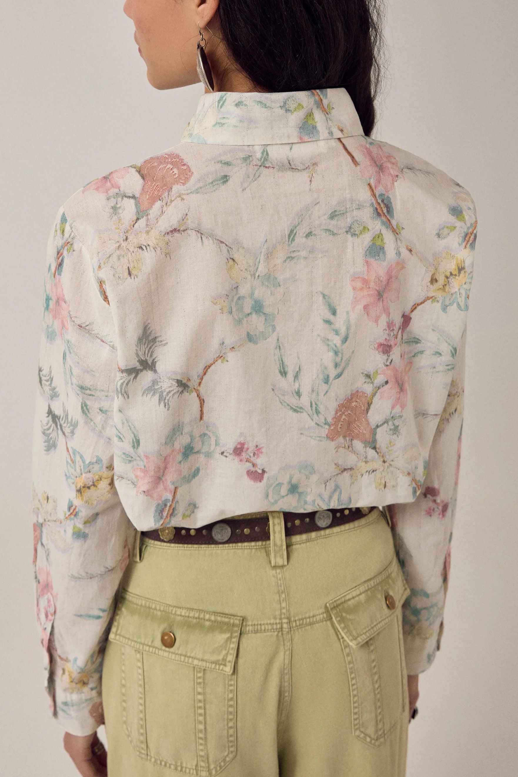 A person faces away, wearing the oversized Narciso Shirt by Maison Hotel—cotton with floral print—tucked into high-waisted pale green pants and a brown belt. Their dark hair is loose, accessorized with a large hoop earring.