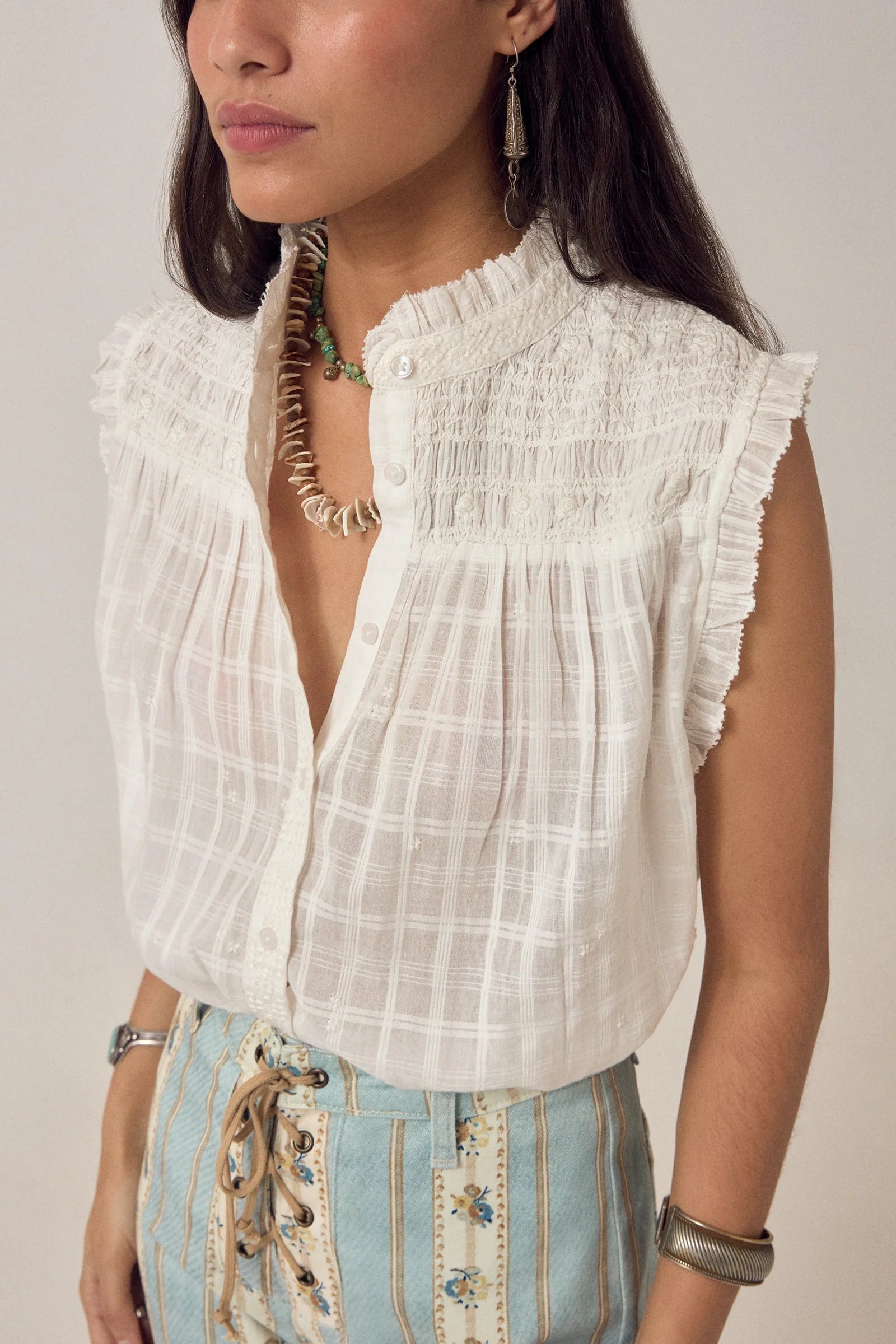 A woman wears the Maison Hotel Elvis Sleeveless Blouse, styled with light blue high-waisted pants featuring embroidery and a lace-up front. She completes the outfit with earrings and layered necklaces.