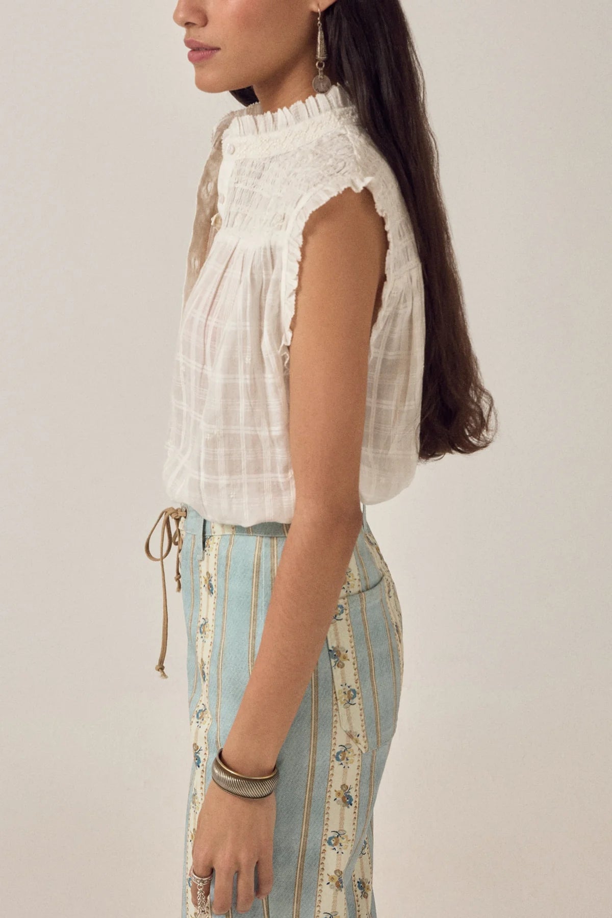 A woman with long brown hair stands in profile against a plain light background, wearing the Maison Hotel Elvis Sleeveless Blouse paired with a high-waisted blue and cream patterned skirt.