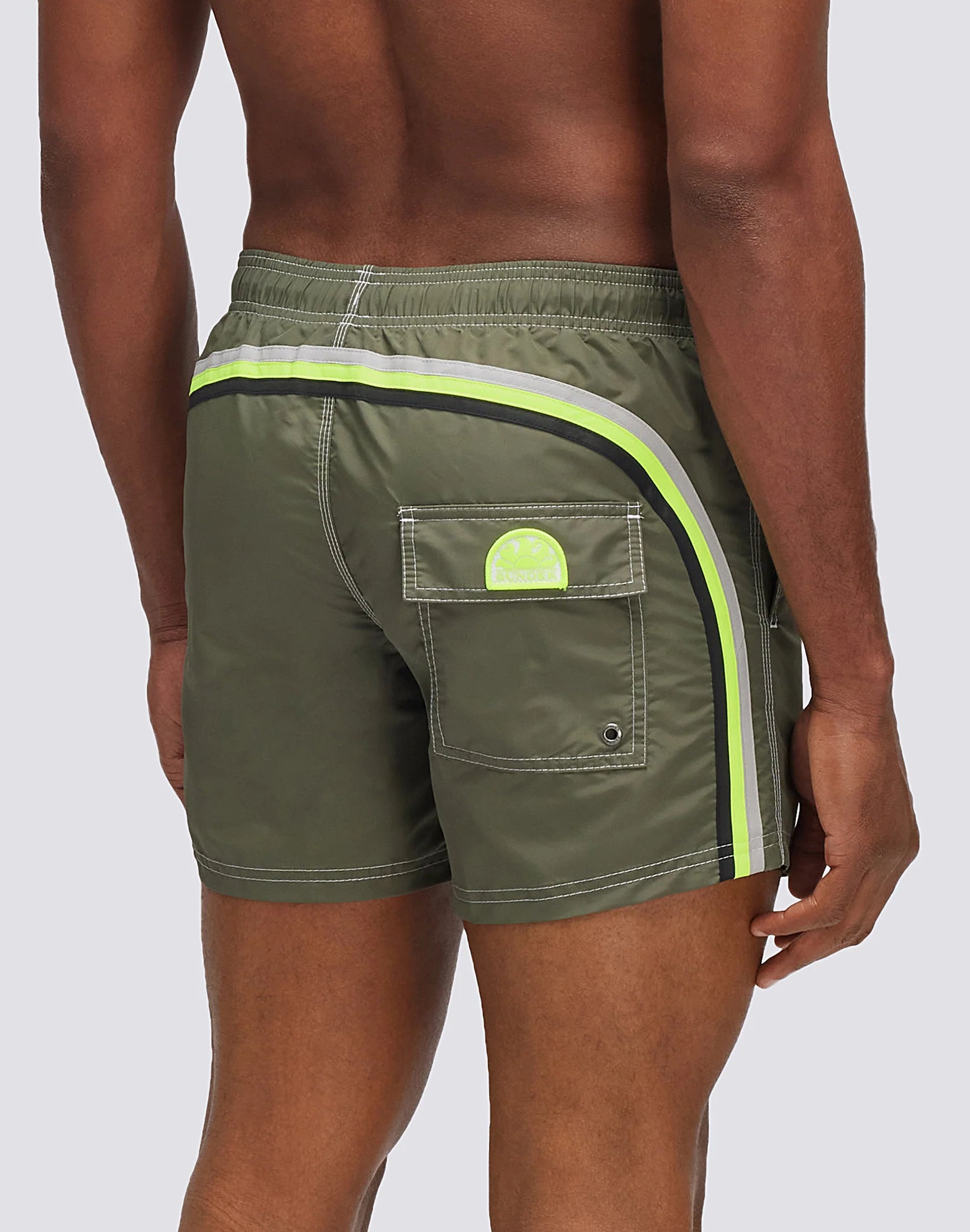A person wearing SUNDEK/KickoffUSA ELASTIC WAIST Board Shorts in olive green nylon-taffeta, with rainbow yellow and black side stripes and a back flap pocket, stands against a light gray background.