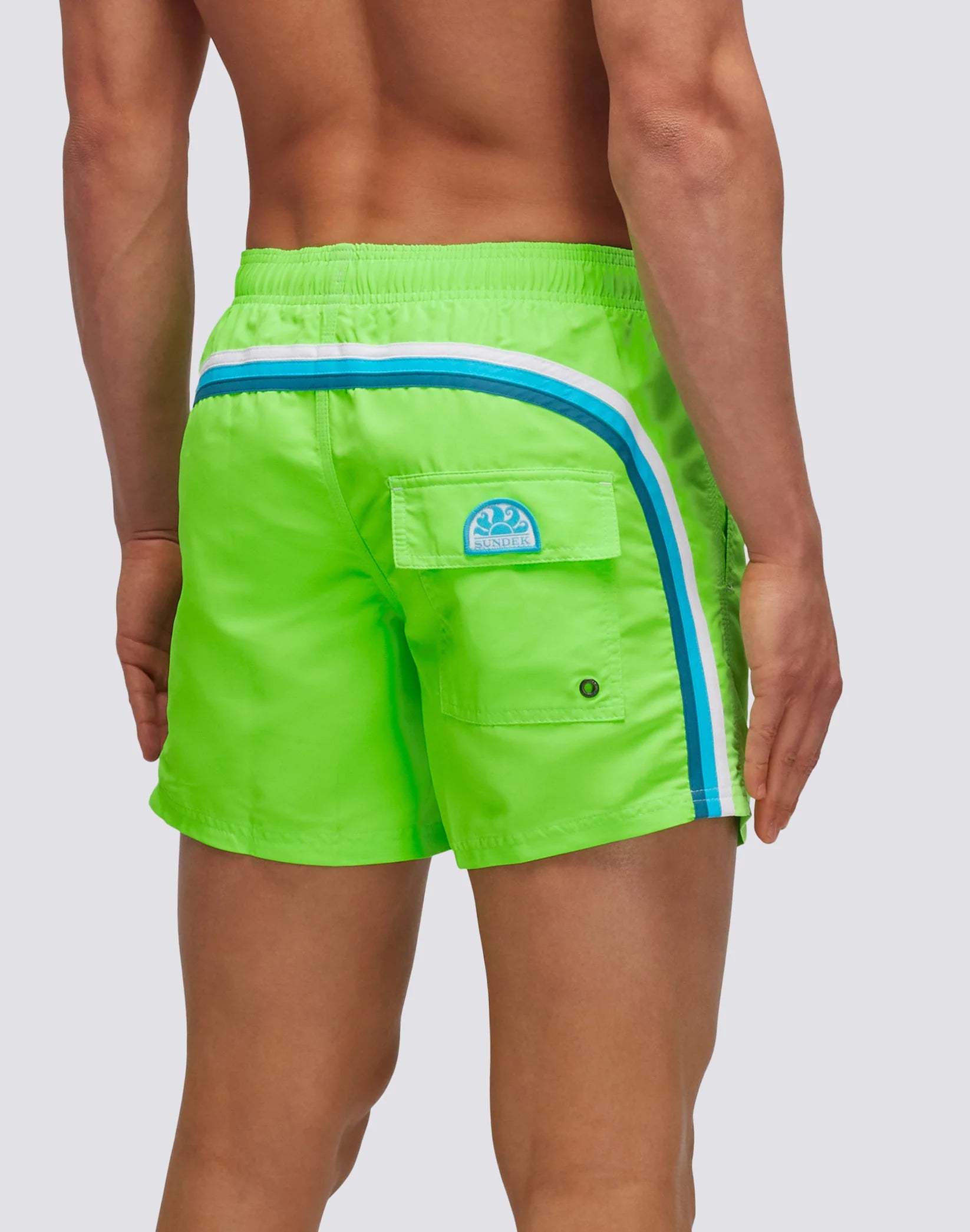A person shown from behind wearing SUNDEK/KickoffUSA ELASTIC WAIST Board Shorts in bright green nylon-taffeta with blue and white side stripes, a flap back pocket with round blue patch logo, and the signature Sundek rainbow.