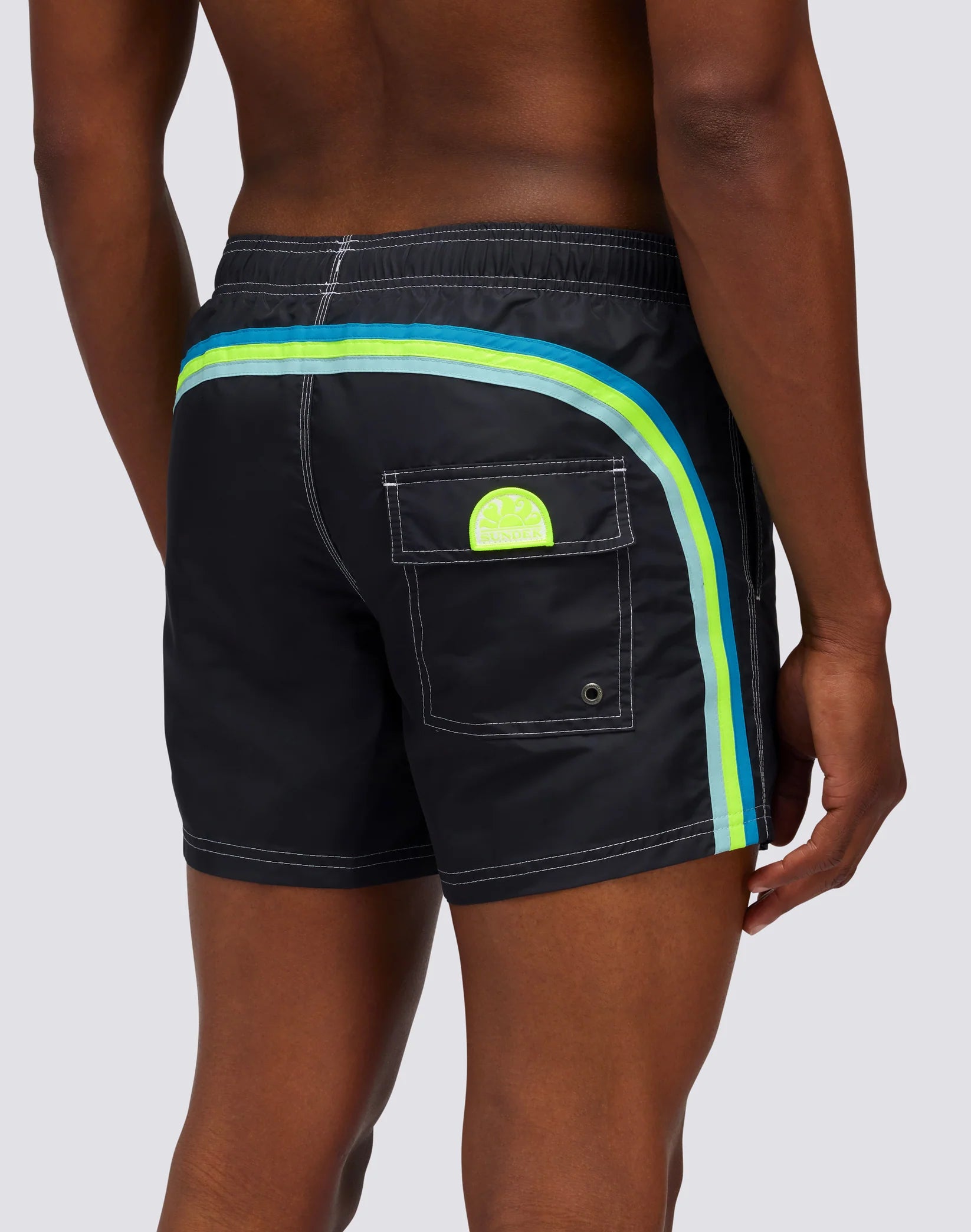 A teen wears SUNDEK/KickoffUSA 14" ELASTIC WAIST Board Shorts—black swim trunks with blue and green side stripes, neon green logo on the back pocket, white stitching, and a comfy elastic waistband.