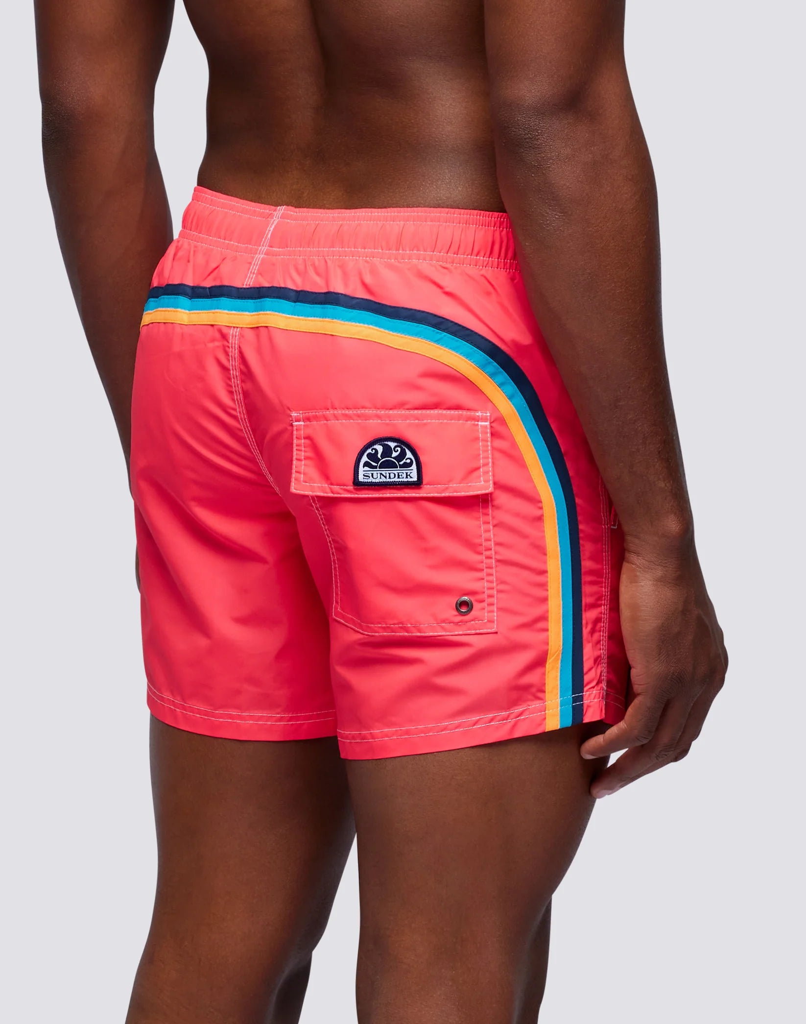 A man models SUNDEK/KickoffUSA 14" ELASTIC WAIST Board Shorts in bright coral with a rainbow side stripe and a back flap pocket, featuring his waist and back against a plain light backdrop.