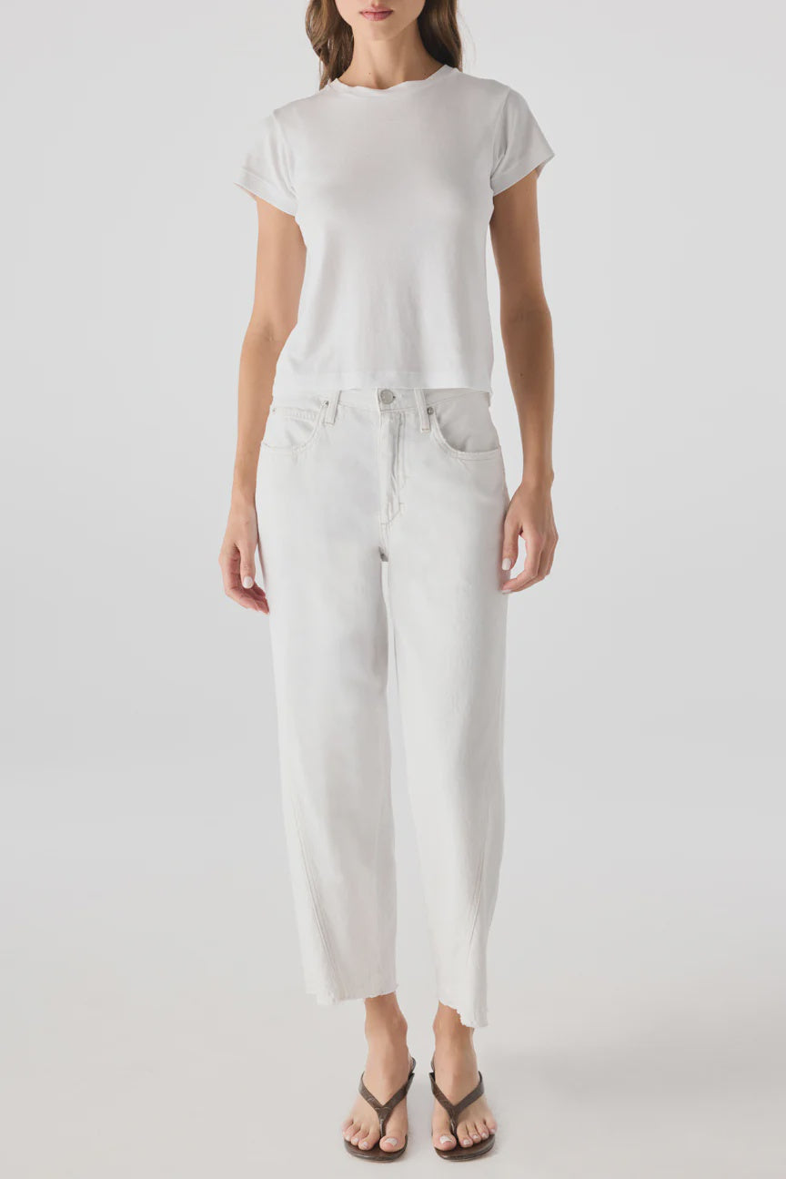 A woman stands facing forward, wearing a plain white short-sleeve t-shirt, AMO Denim's Maria Denim vintage-inspired high-waisted white jeans with a loose fit, and black flip-flops. The background is light and plain.