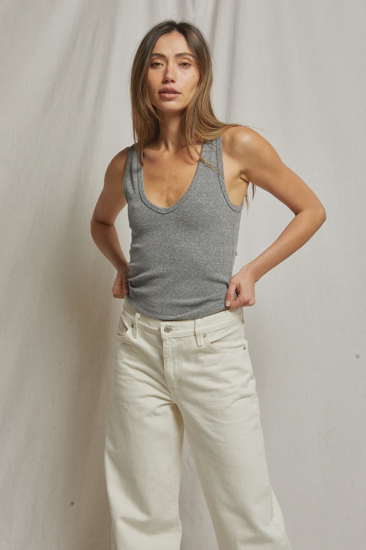 A woman with long brown hair stands against a neutral backdrop, wearing the Maria Rib V Tank by Perfectwhitetee and white pants. She looks at the camera with a relaxed expression, her hands tucked into her waistband.