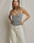 A woman with long brown hair stands against a neutral backdrop, wearing the Maria Rib V Tank by Perfectwhitetee and white pants. She looks at the camera with a relaxed expression, her hands tucked into her waistband.