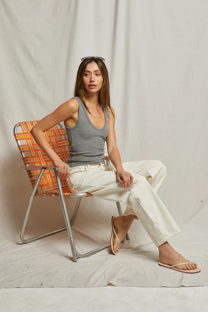 A woman sits on an orange and white checkered chair, wearing the Maria Rib V Tank by Perfectwhitetee in gray, paired with white pants and brown sandals. Sunglasses rest on her head, against a plain light-colored fabric background.