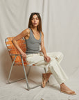 A woman sits on an orange and white checkered chair, wearing the Maria Rib V Tank by Perfectwhitetee in gray, paired with white pants and brown sandals. Sunglasses rest on her head, against a plain light-colored fabric background.