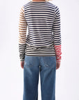 An individual with blonde hair faces away from the camera, donning blue jeans and the AMO Denim Marla Tee, a chic high-necked, long-sleeve shirt made of 100% cotton. It features stylish tri-color stripes in black, white, and red.