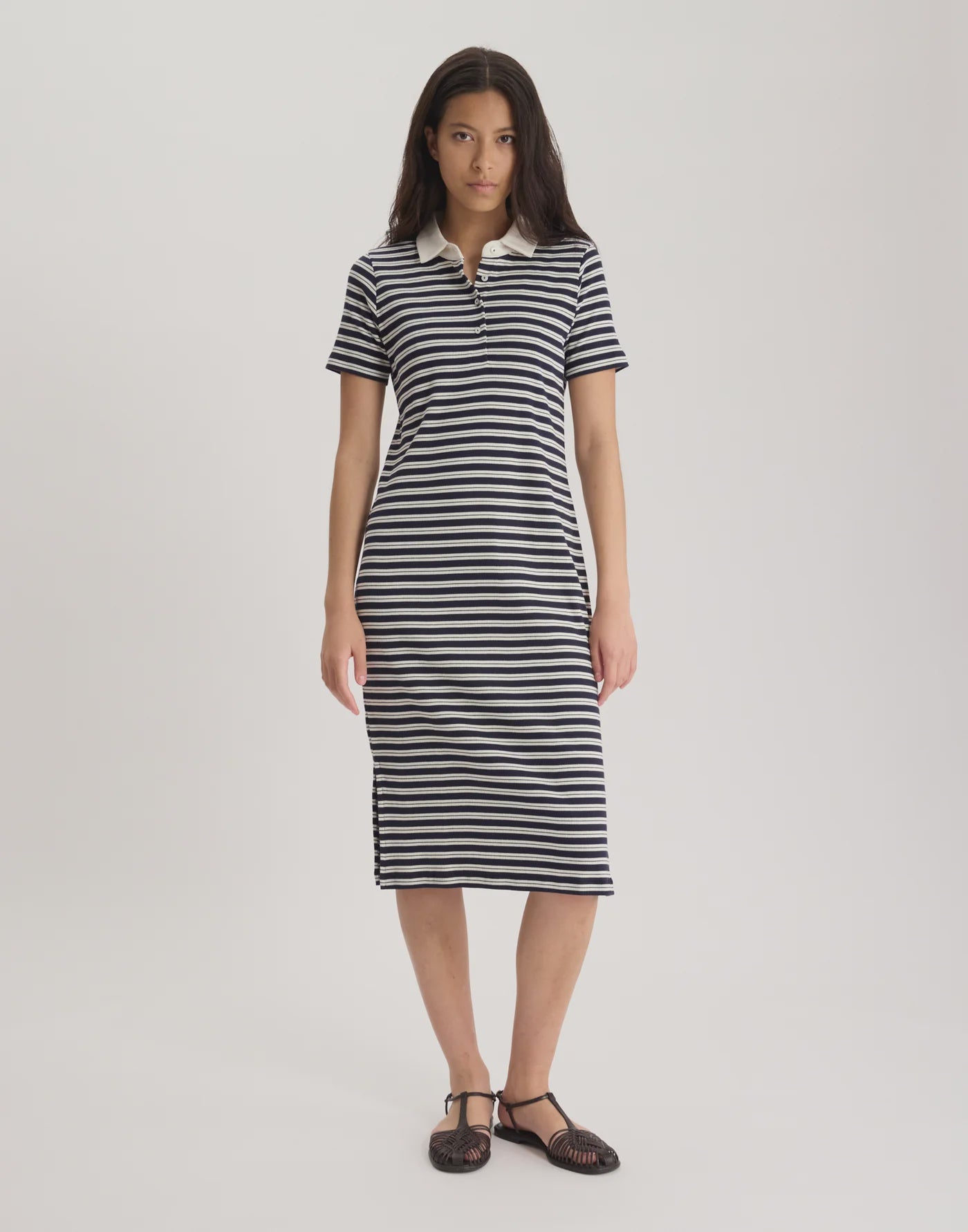 A woman poses against a plain background wearing the black-and-white striped, organic cotton Romane Polo Dress by Ines De La Fressange, featuring a white collar and midi length, paired with black shoes.