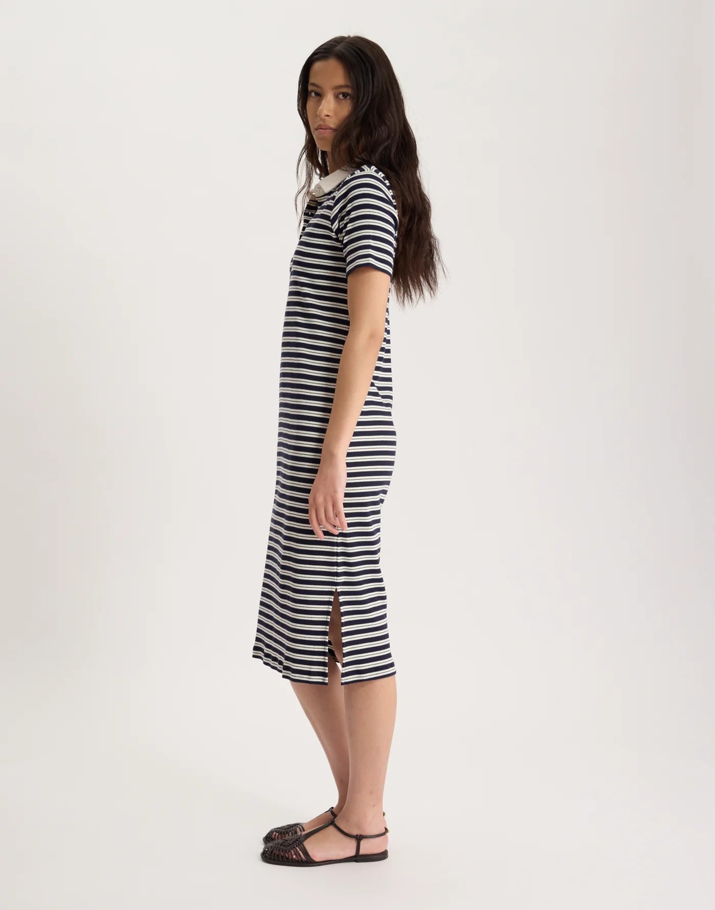 A woman with long dark hair stands sideways in front of a plain light background, wearing the Ines De LA Fressange Romane Polo Dress—an organic cotton black and white striped midi dress with a side slit—and black sandals.