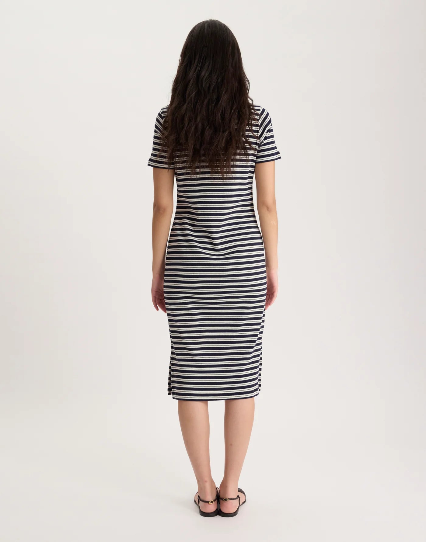 A woman with long, wavy brown hair stands facing away in the Ines De LA Fressange Romane Polo Dress—an organic cotton black and white striped midi dress—with black sandals, set against a plain white background.