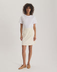 A person with curly hair stands facing forward in a plain white t-shirt and the Ines De LA Fressange Summer Skirt, a cream-colored elastic waist skirt with front pockets, paired with tan lace-up shoes against a plain white background.