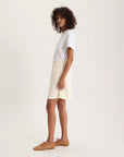 A person with curly hair stands sideways against a light background, wearing a white t-shirt and the Ines De LA Fressange Summer Skirt—an off-white terry cotton skirt with front pockets and an elastic waist—paired with tan flat shoes.