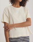 A person wears an off-white, short-sleeved Naomi Tee Shirt by Ines De LA Fressange in terry cloth with a small oak leaf logo, paired with a navy and white striped skirt. The person’s face is partially out of frame.