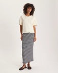 A person with curly hair wears an Ines De LA Fressange Naomi Tee Shirt made from 100% cotton and a long black-and-white striped skirt, paired with black sandals, standing against a plain light background.