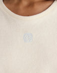 Close-up of a person wearing the cream-colored Naomi Tee Shirt by Ines De LA Fressange, featuring terry cloth fabric and a small light blue embroidered emblem centered below the round neckline.