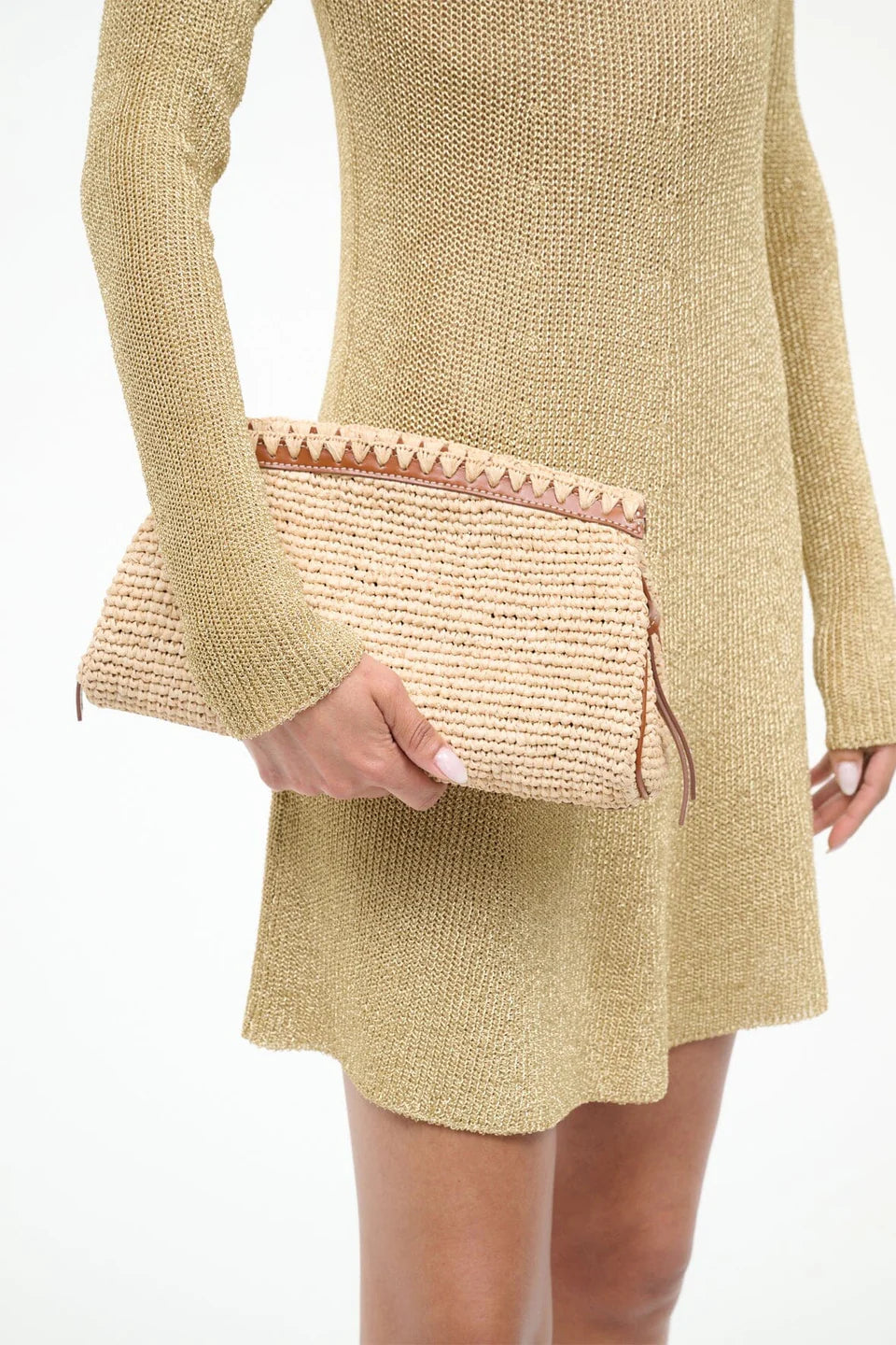 A person in a gold knit dress holds the Staud Maude Convertible Clutch in Natural, featuring raffia with brown leather trim, zipper closure, and a removable leather strap, against a plain white background.