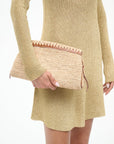 A person in a gold knit dress holds the Staud Maude Convertible Clutch in Natural, featuring raffia with brown leather trim, zipper closure, and a removable leather strap, against a plain white background.