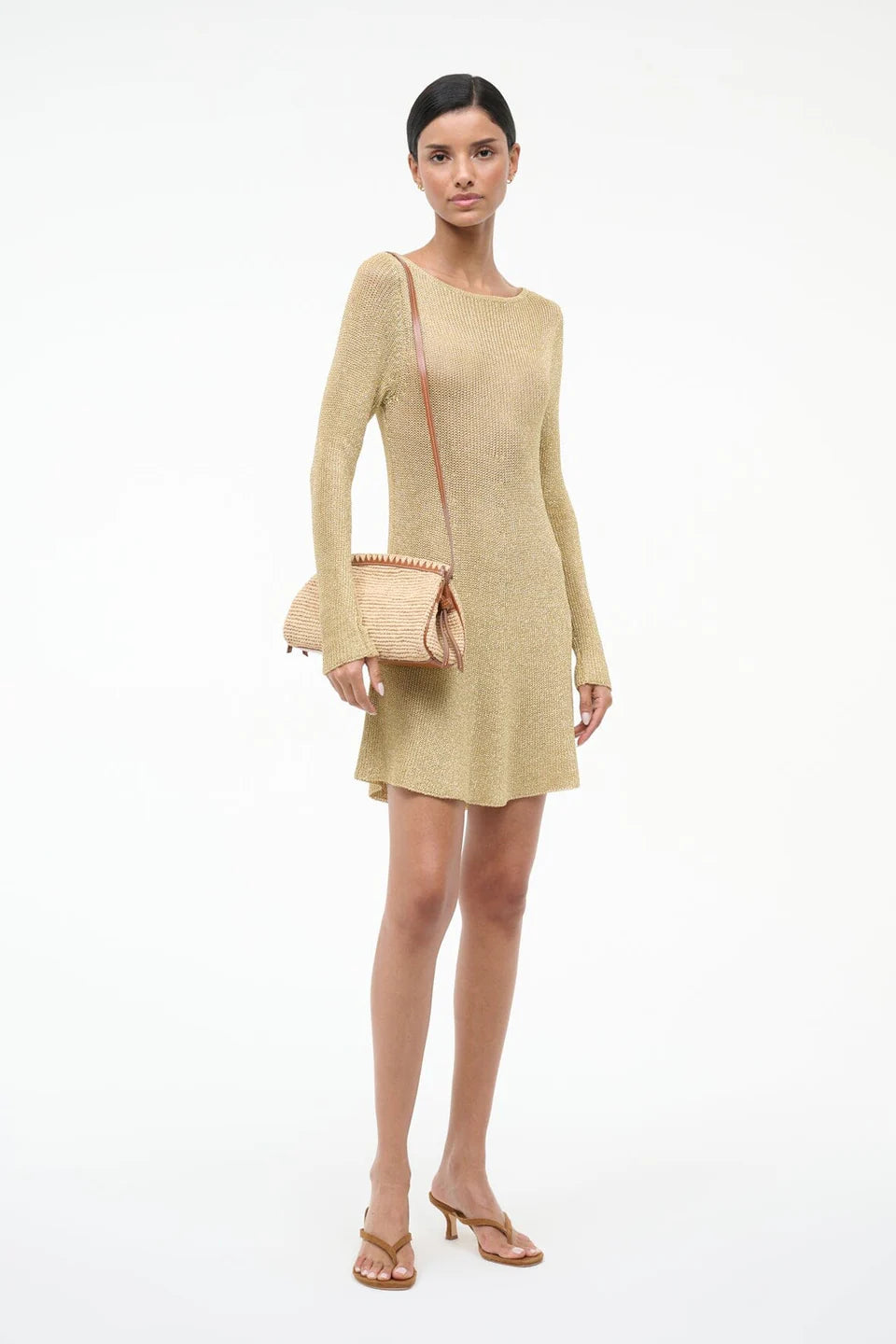 A woman poses against a white background wearing a long-sleeve gold mini dress, tan heeled sandals, and the Staud Maude Convertible Clutch in Natural, holding its removable leather strap in one hand.