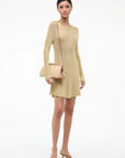 A woman poses against a white background wearing a long-sleeve gold mini dress, tan heeled sandals, and the Staud Maude Convertible Clutch in Natural, holding its removable leather strap in one hand.