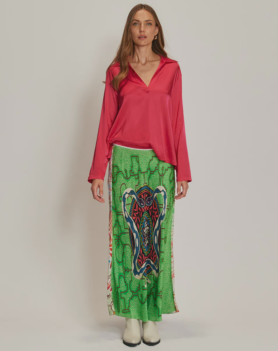 A woman with long brown hair wears a bright pink long-sleeved blouse, white boots, and the Me369 Blakely Slip Skirt—a green maxi skirt featuring a bold abstract pattern in red, blue, and black.