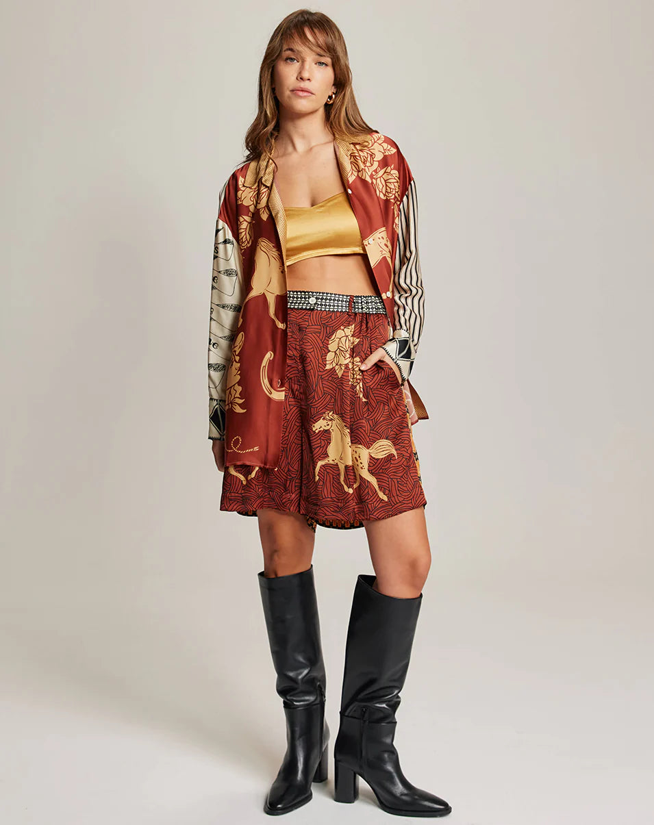 A woman wears the Me369 Juliette Pleated Shorts—a red and gold patterned set with an elastic waist—paired with a yellow crop top, checked belt, black knee-high boots, and loose brown hair as she faces the camera neutrally.